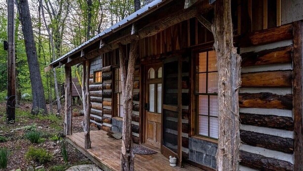 Una cabaña rústica de troncos con un porche de madera, ventanas amplias y cimientos de piedra.
