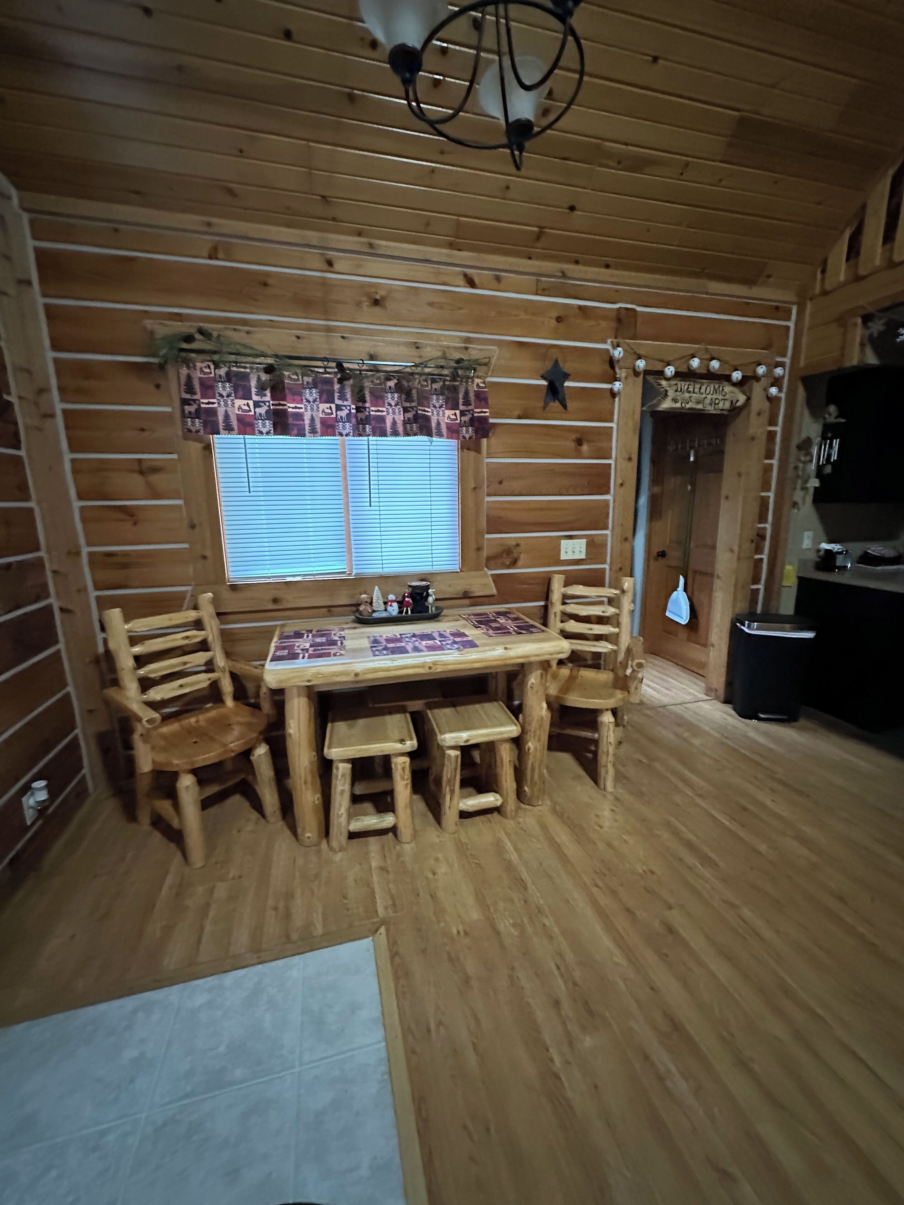 Un intérieur en bois avec une table et des chaises, une fenêtre avec des stores et une pancarte « Bienvenue ».