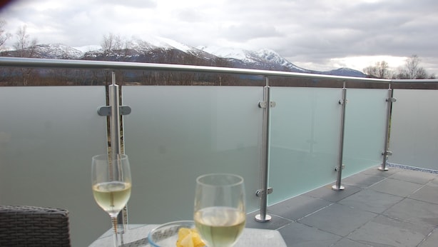 A balcony with a glass railing, two wine glasses, a bowl of fruit, and a wicker chair. - Glas Beag (Spean Bridge)