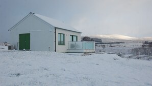 A small, single-story building with a green door and windows, surrounded by snow-covered fields and distant hills. - Glas Beag (Spean Bridge)
