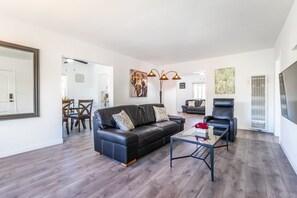 Uma sala de estar moderna com um sofá de couro preto, uma mesa de centro de vidro e uma poltrona de couro preto. Ao fundo, uma área de jantar com cadeiras de madeira e uma mesa.
