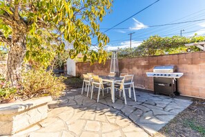 Um pátio externo com mesa e cadeiras de pedra, churrasqueira e uma árvore.