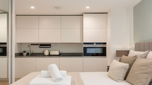 A modern bedroom with a large bed, a bedside table, and a built-in kitchen area. - The Kingston Upon Thames Apartme (Kingston Upon Thames)