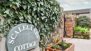Opastekyltti, jossa lukee ”Scaleland’s Cottage”, ja taustalla näkyy murattiseinillä varustettu mökki sekä tiilestä rakennettu rakennelma.