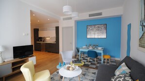 A modern living room with a sofa, a TV, a dining area with a table and chairs, and a kitchen in the background.