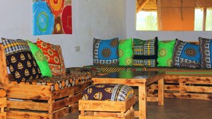A room with a wooden sofa set and a matching coffee table, a colorful abstract painting on the wall, and a window with a view of trees. - Nguge Hills Lodge & Wellness Retreat (Nyamboyo)