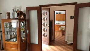 Een kamer met een houten kast vol glaswerk en servies, en een deur die leidt naar een keuken met houten kasten en een witte koelkast.