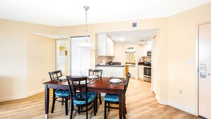 A dining area with a wooden table and chairs, a kitchen in the background, and a door to the right.