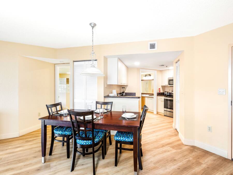 A dining area with a wooden table and chairs, a kitchen in the background, and a door to the right.
