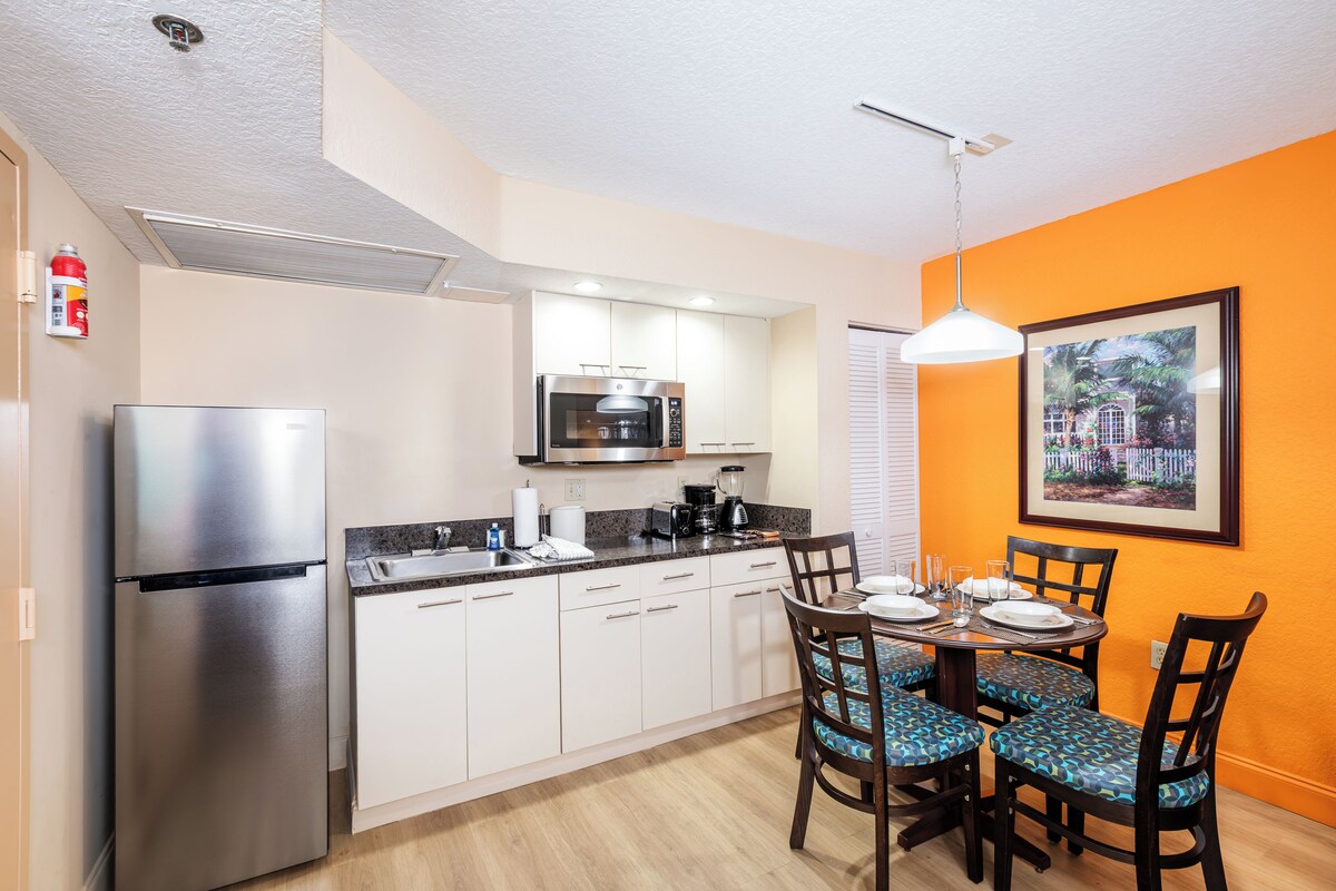 A compact kitchen with a refrigerator, microwave, and coffee maker. There is a dining table with chairs and a framed picture on the wall.