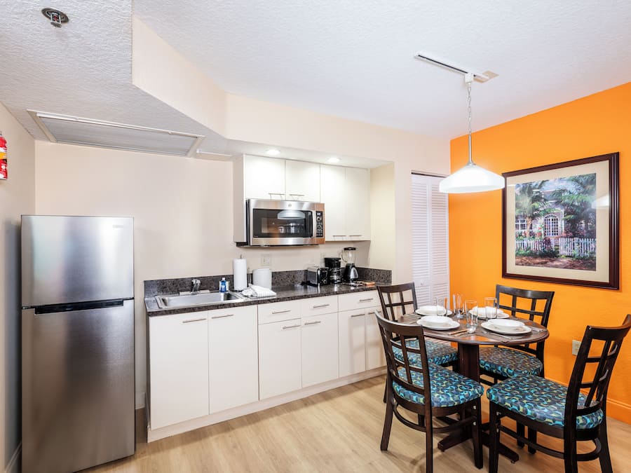A compact kitchen with a refrigerator, microwave, and coffee maker. There is a dining table with chairs and a framed picture on the wall.