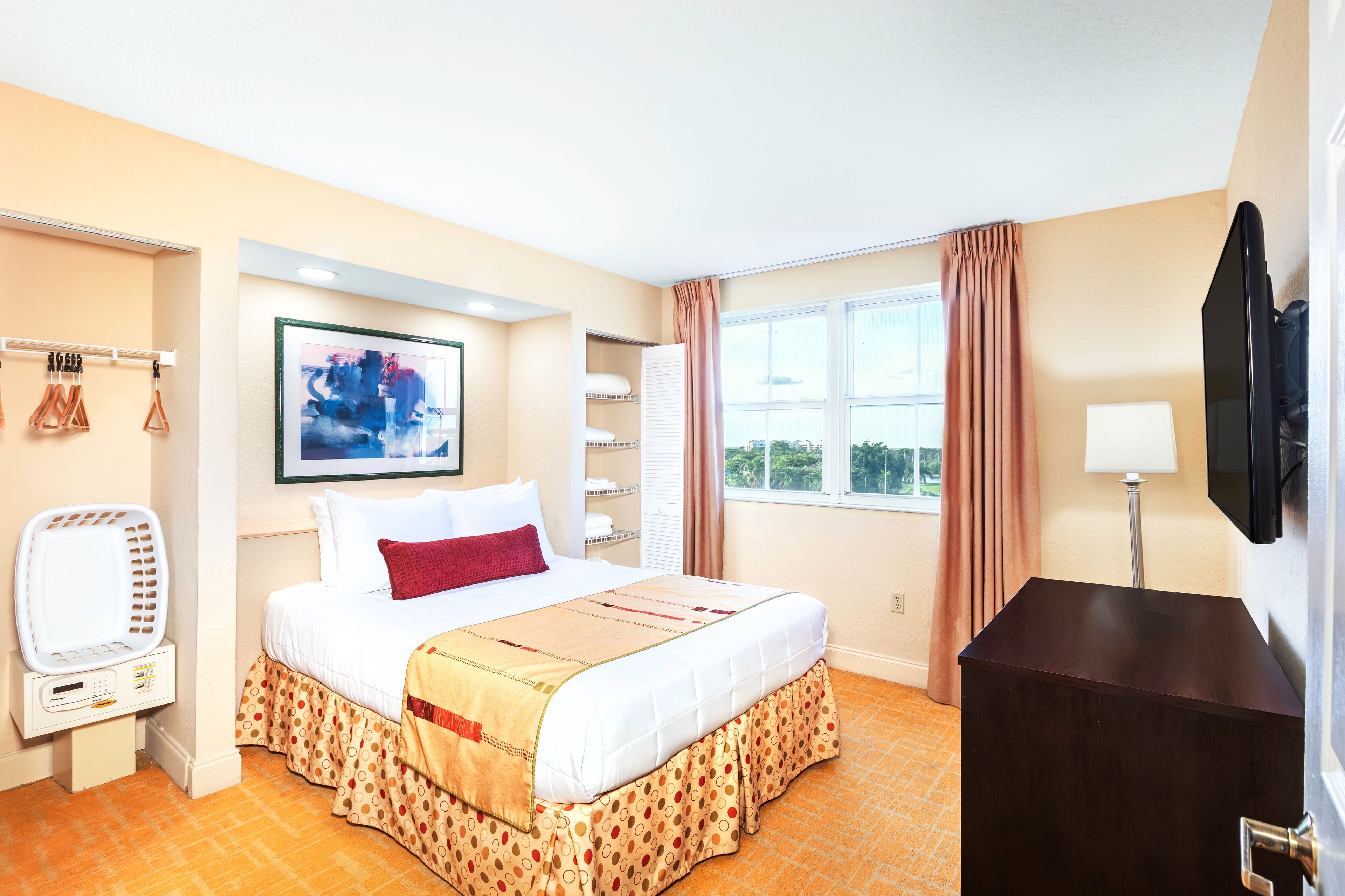 Une chambre d’hôtel avec un lit, une télévision, un bureau, une fenêtre avec des rideaux et un tableau au mur.