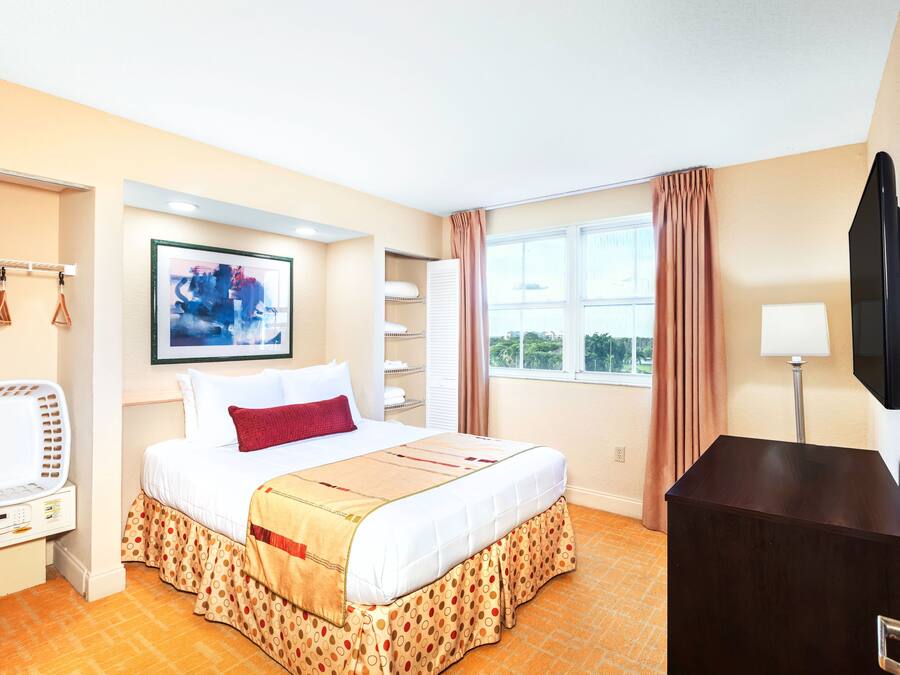 A hotel room with a bed, a TV, a desk, a window with curtains, and a painting on the wall.