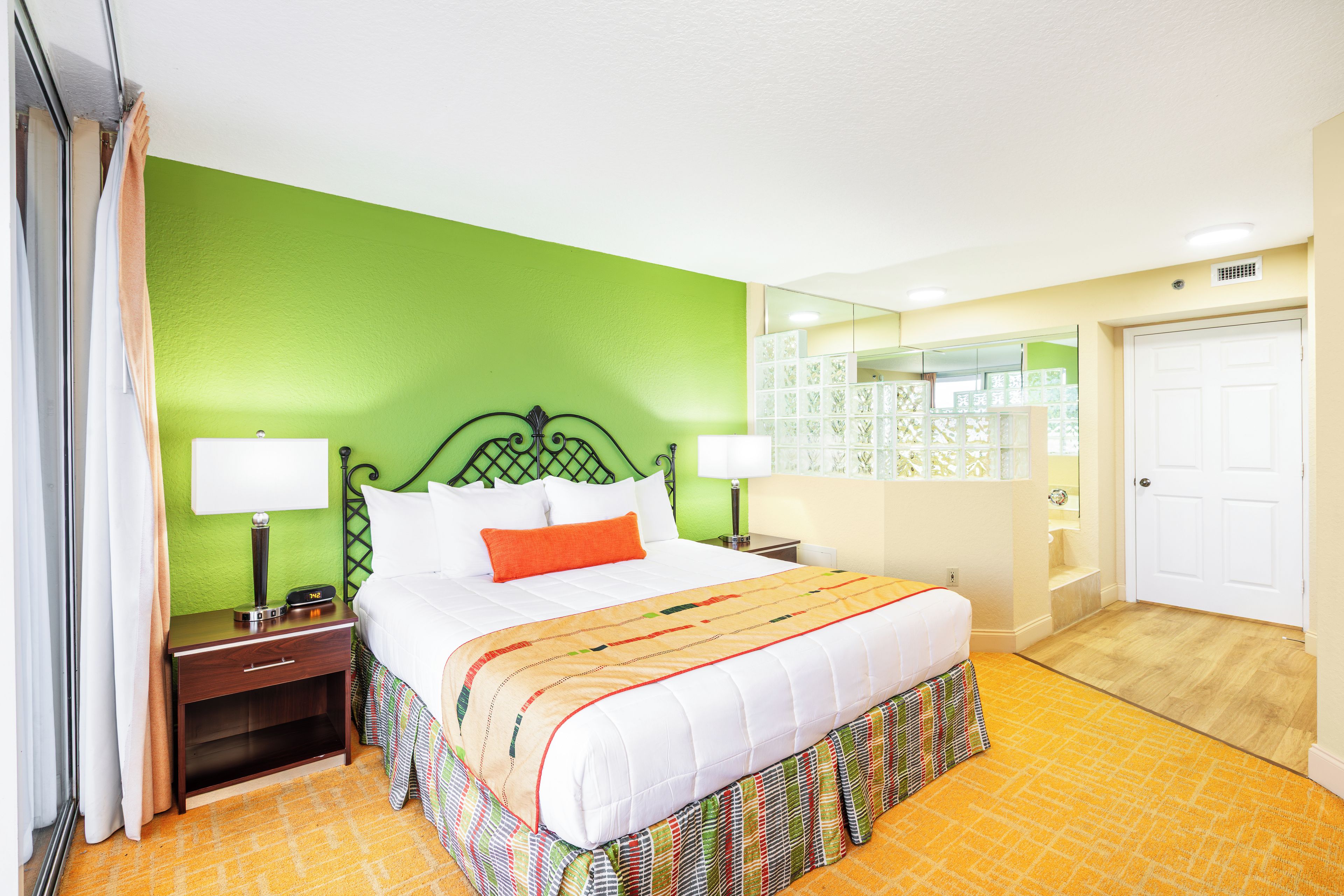 Une chambre d’hôtel avec un mur vert, un lit blanc avec un oreiller orange, deux tables de chevet avec des lampes, un miroir et une porte.
