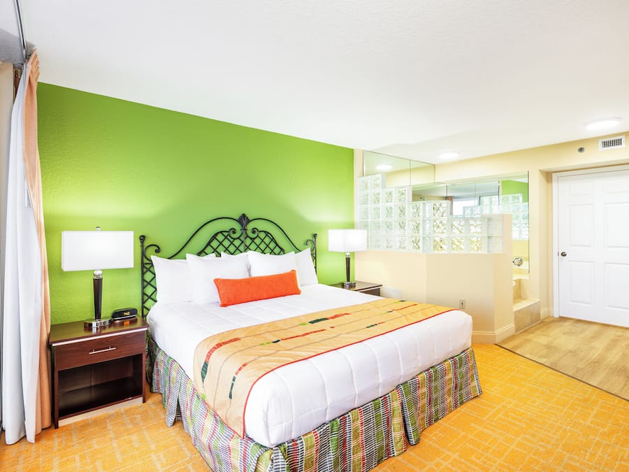 A hotel room with a green wall, a white bed with an orange pillow, two bedside tables with lamps, a mirror, and a door.