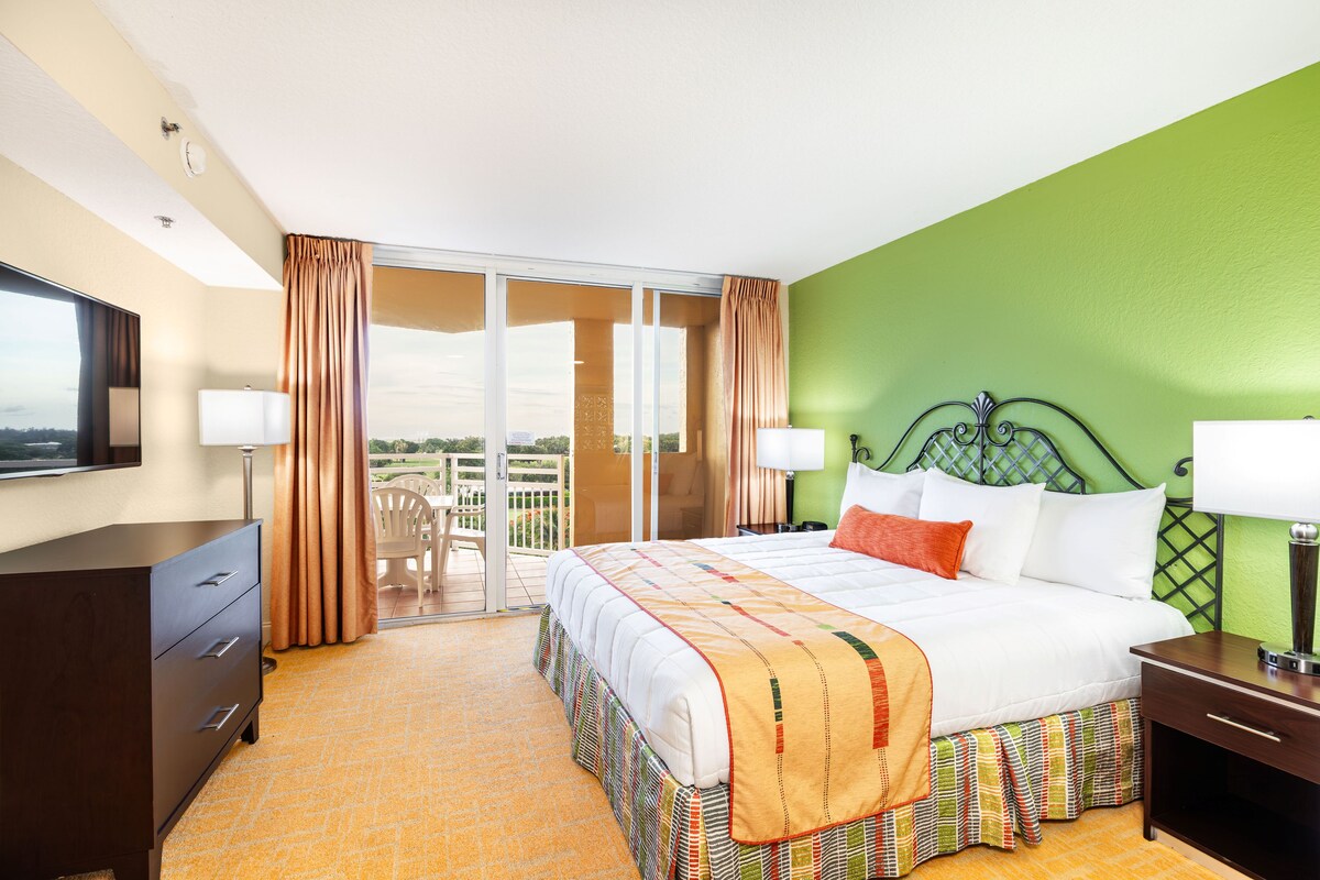 A hotel room with a bed, a desk, a chair, a TV, and a balcony with curtains.