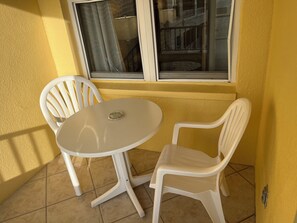 A small outdoor seating area with a round table and two white plastic chairs, a window with curtains, and a yellow wall.
