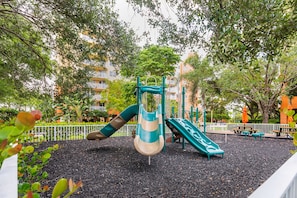 A playground with slides and climbing structures, surrounded by trees and a fence. - Vacation Village at Bonaventure (Weston)