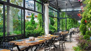 Un café al aire libre con mesas y sillas, amplias ventanas y abundante vegetación.