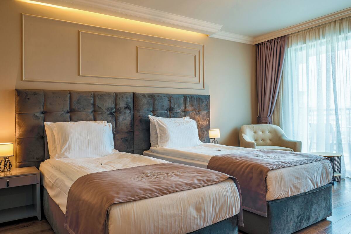 A hotel room with two beds, a large padded headboard, a chair, and a large window with curtains.