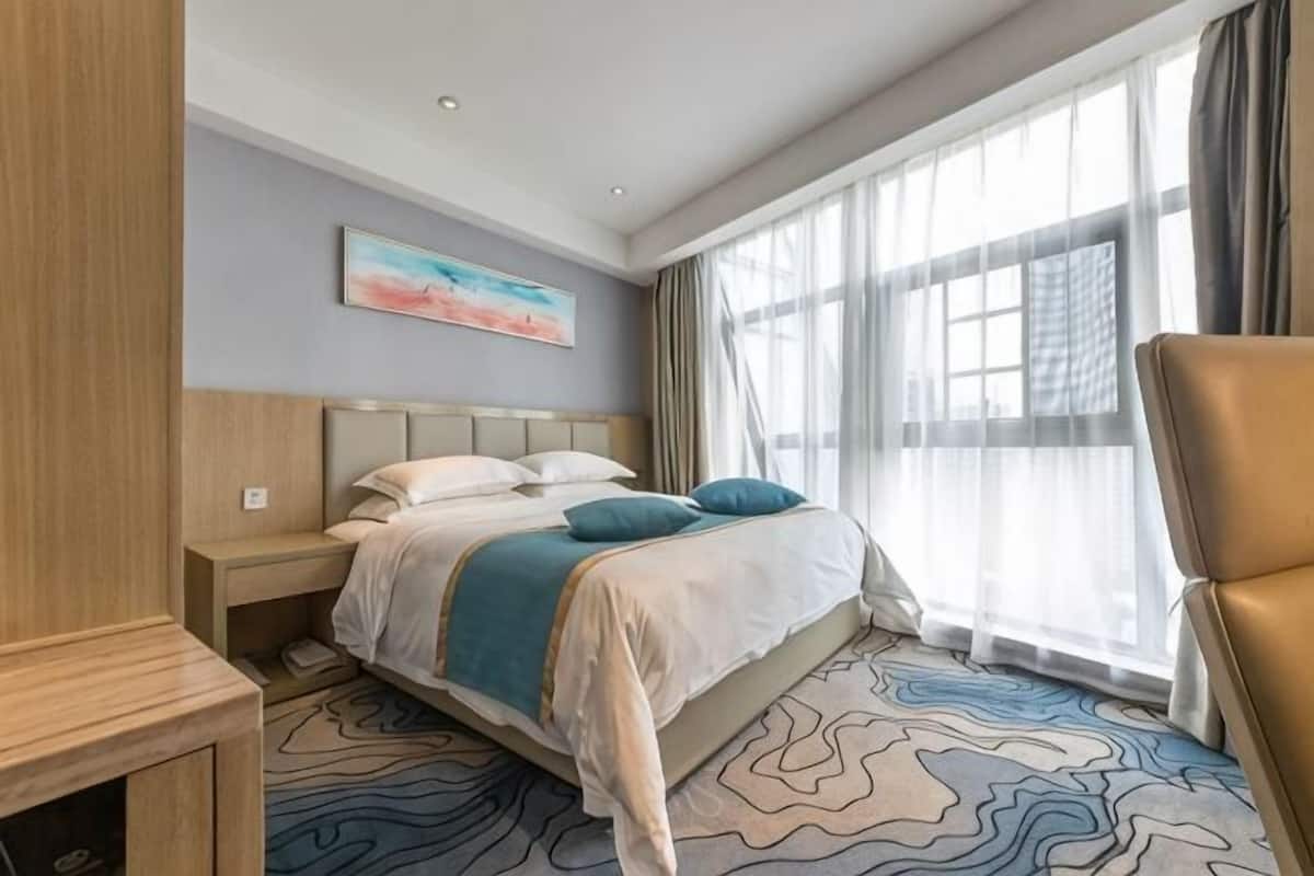 Una habitació d'hotel amb llit, una cadira, una tauleta de nit, un quadre a la paret i una finestra amb cortines.