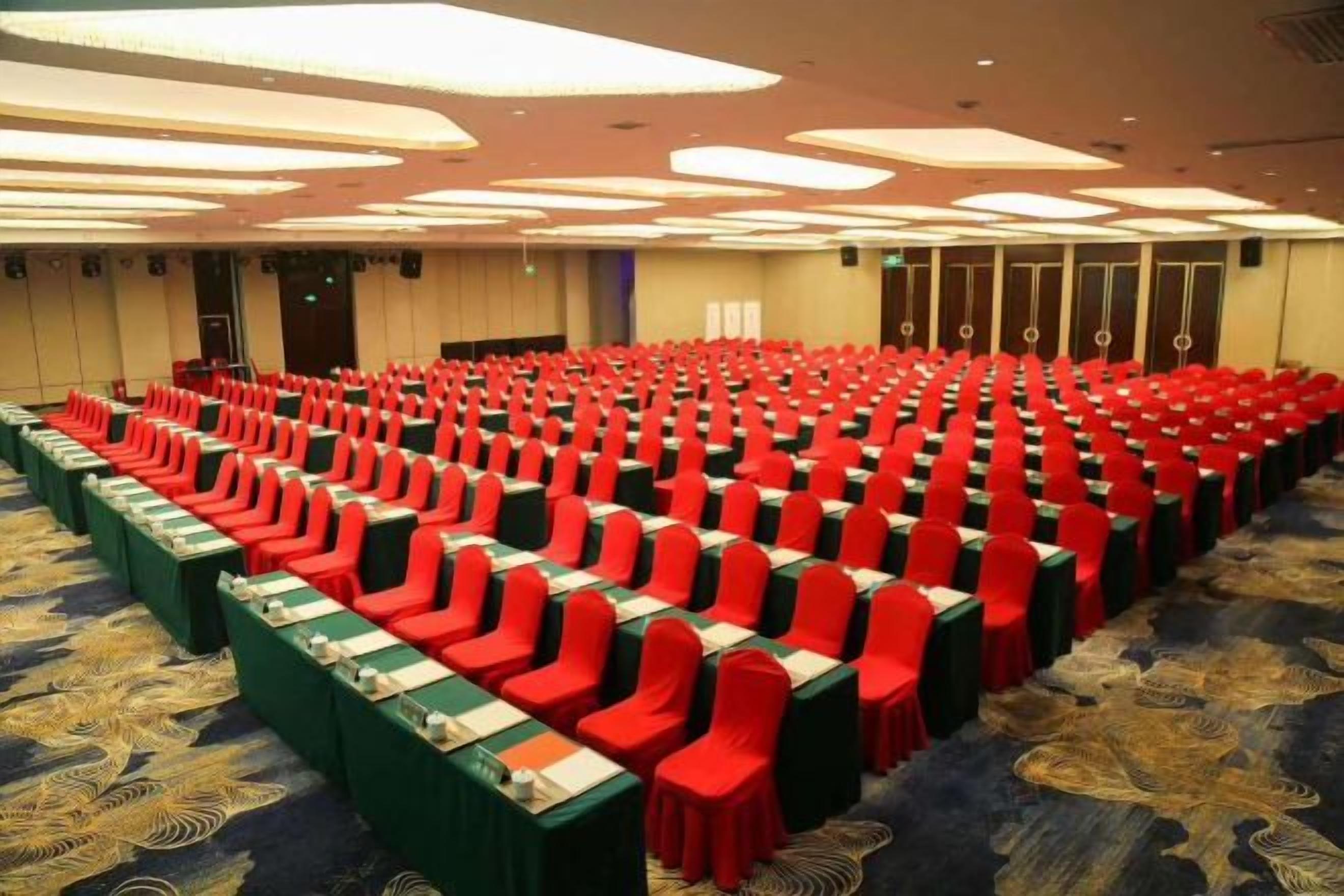 Une salle de conférence avec des rangées de chaises et de tables, un sol recouvert de moquette et un plafond doté d’un éclairage encastré.