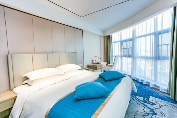 A hotel room with a large bed, a nightstand, a desk, and a view of the outdoors through large windows. - Donghuang International Hotel (Xinhua)
