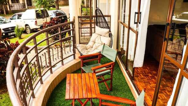 A balcony with a wooden table and chairs, a hanging chair, and a view of parked cars and greenery.