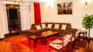 A living room with a brown sofa, wooden coffee table, and patterned armchairs. There is a red rug, a potted plant, and a painting on the wall.
