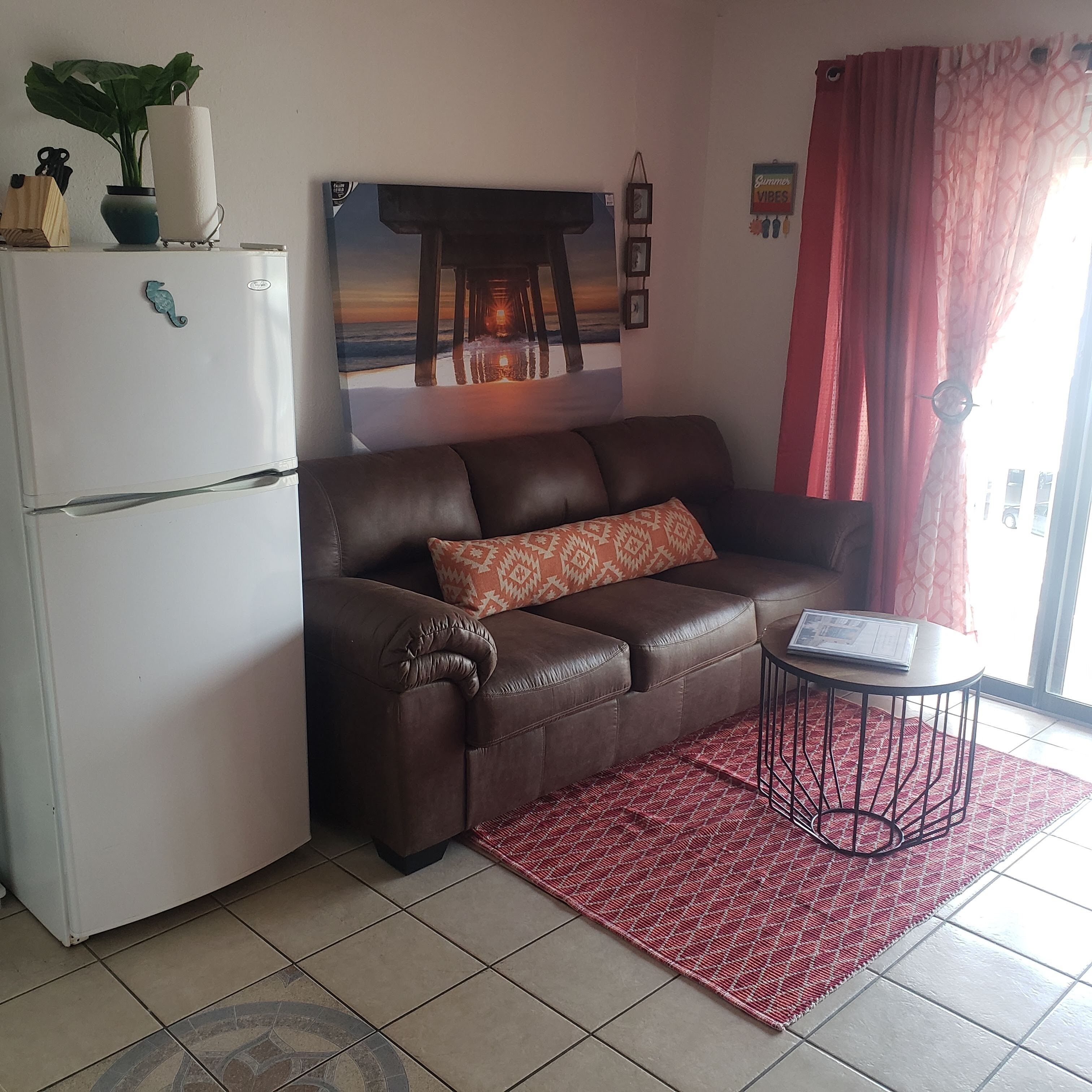 Een woonkamer met een bruine leren bank, een witte koelkast, een rood tapijt met een patroon en een schilderij van een pier aan de muur.