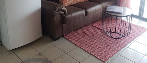 Een woonkamer met een bruine leren bank, een witte koelkast, een rood tapijt met een patroon en een schilderij van een pier aan de muur.