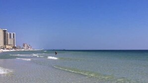 Een strand met helder ondiep water, een persoon die surft, en gebouwen op de achtergrond.