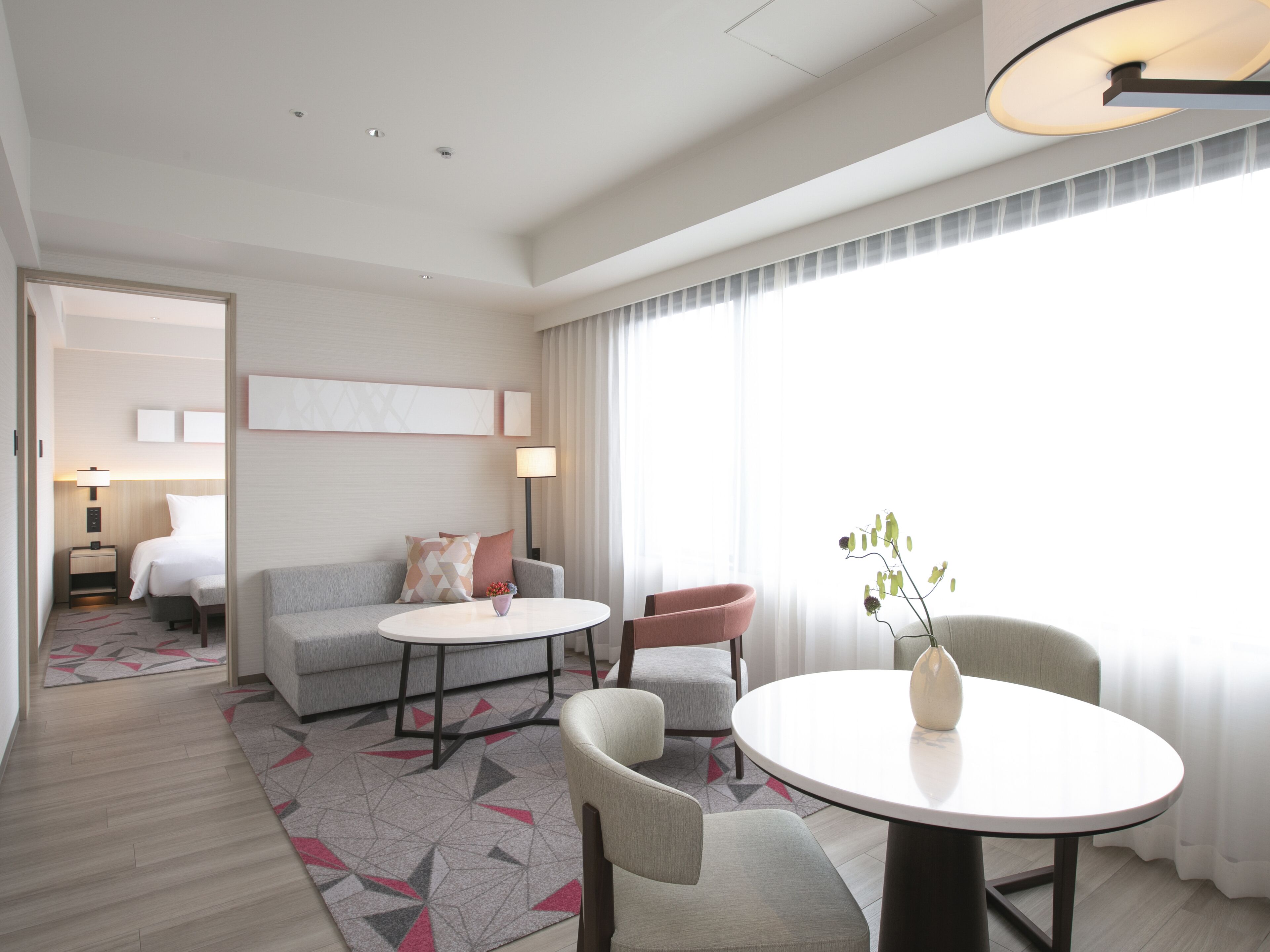 Una habitación de hotel moderna con un sofá gris, una mesa redonda y una silla. Se ve una cama al fondo y una ventana con cortinas.