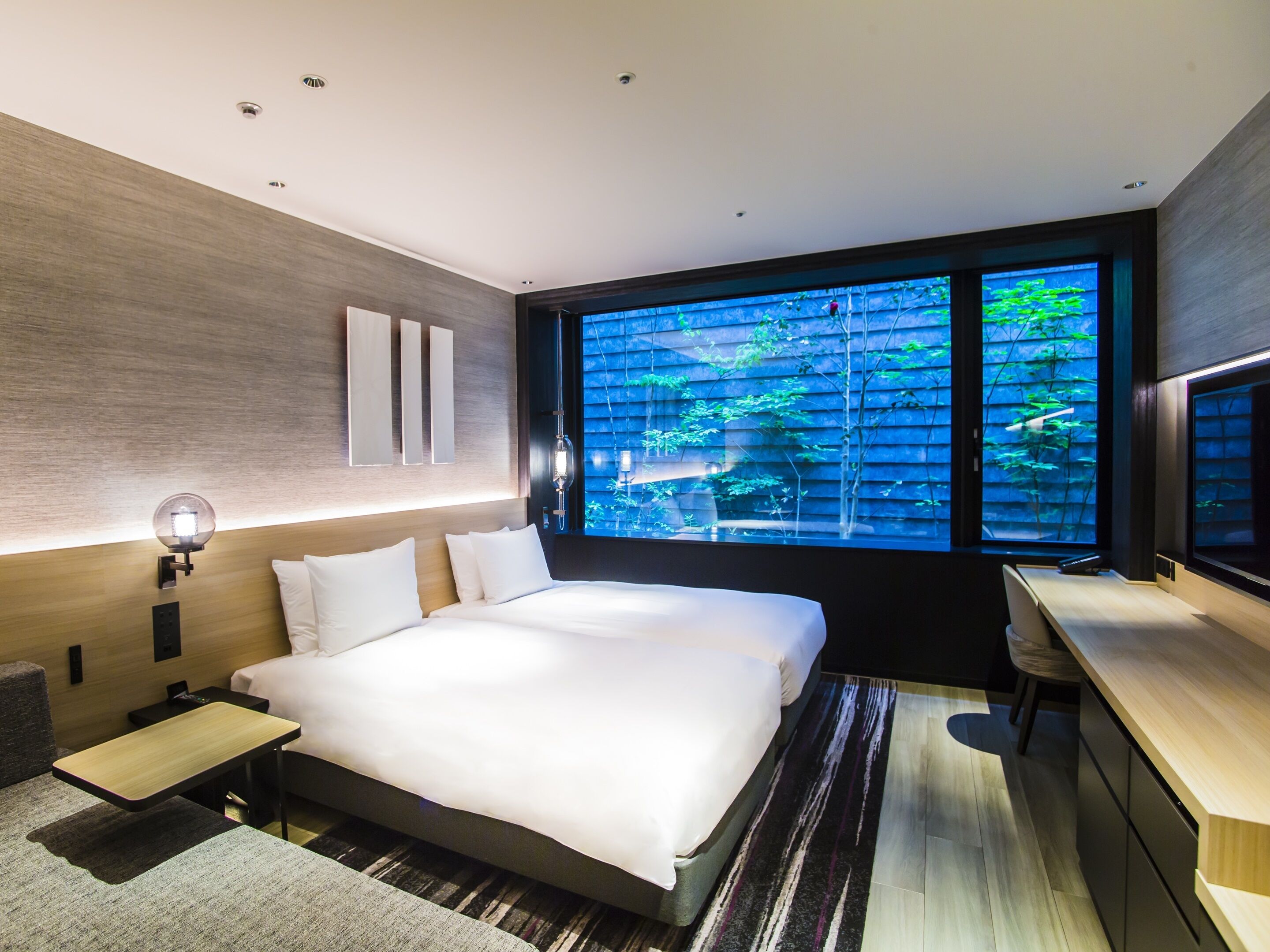 A modern hotel room with a large bed, bedside tables, a desk, and a large window with a view of greenery.