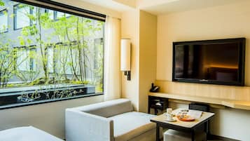 Une chambre d’hôtel avec un lit, un canapé, une petite table avec une assiette et une tasse, un téléviseur fixé au mur et une fenêtre donnant sur des arbres et des immeubles.