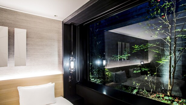 Una habitación de hotel moderna con un ventanal grande, una cama con ropa de cama blanca y un aplique de pared.