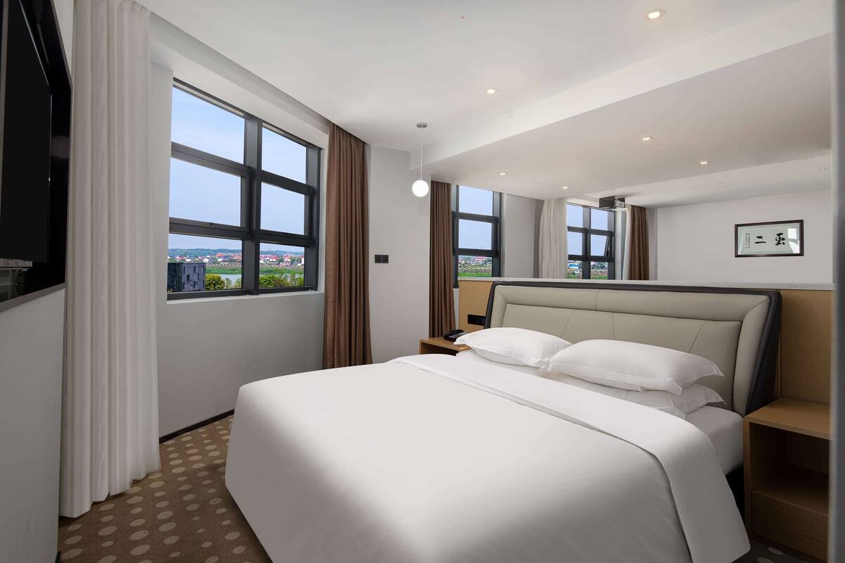 Une chambre d’hôtel avec un grand lit, une table de chevet, une commode et une vue sur la ville grâce à de grandes fenêtres.