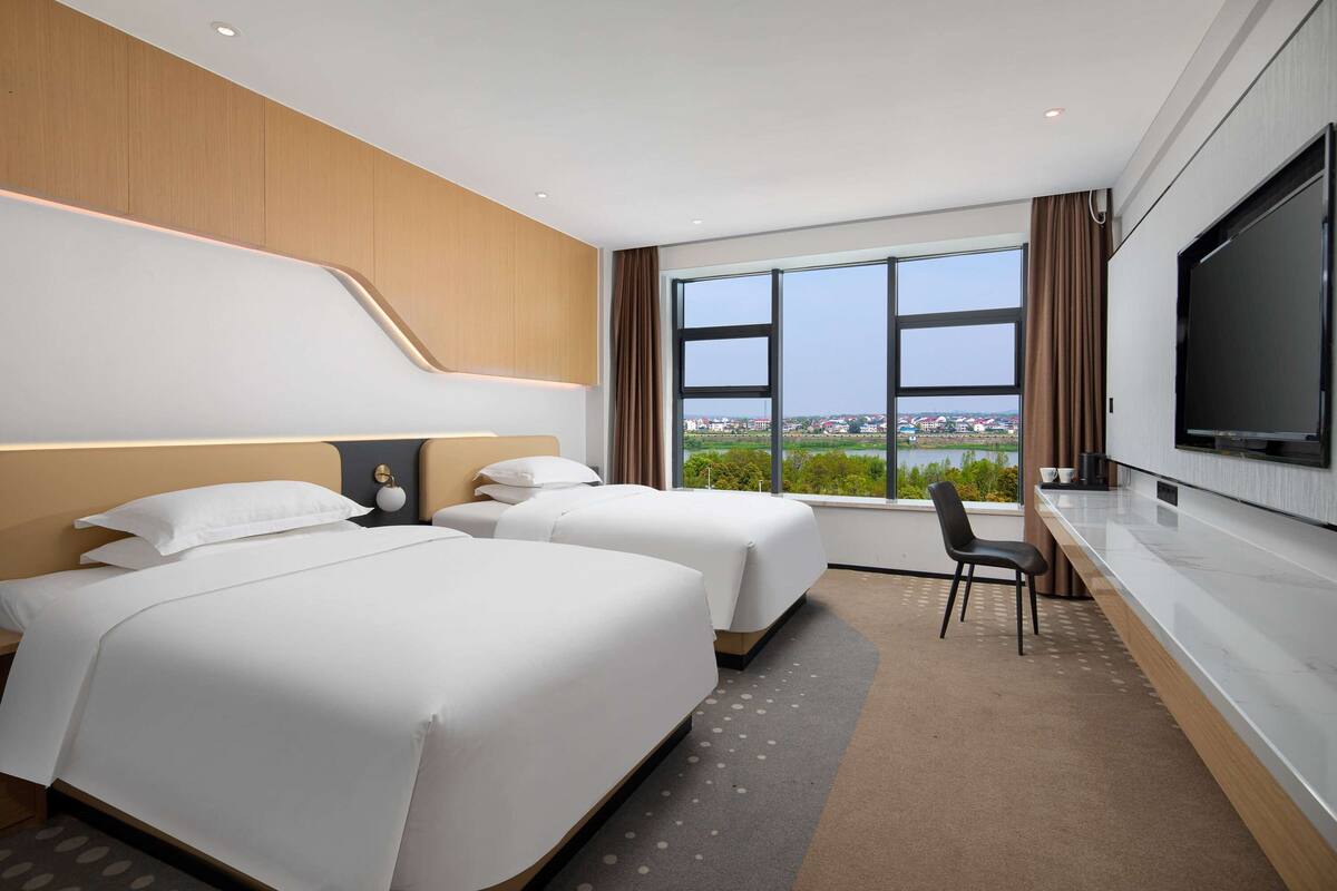 Une chambre d’hôtel avec deux lits, une télévision, un bureau et une grande fenêtre donnant sur un beau paysage.