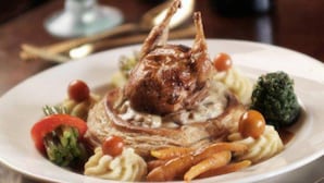 Une assiette de nourriture comprenant un canard rôti, des légumes et une bouteille de vin en arrière-plan.