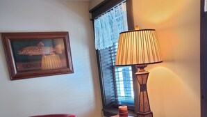 A room with a red leather chair, a wooden table with a lamp, a framed picture on the wall, and a window with lace curtains. - LOVELY/CENTRALLY LOCATED/GROUND FLOOR APARTMENT (Warrensburg)