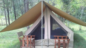 Uma tenda com duas cadeiras de madeira e uma mesa, montada em uma área gramada com árvores ao fundo.