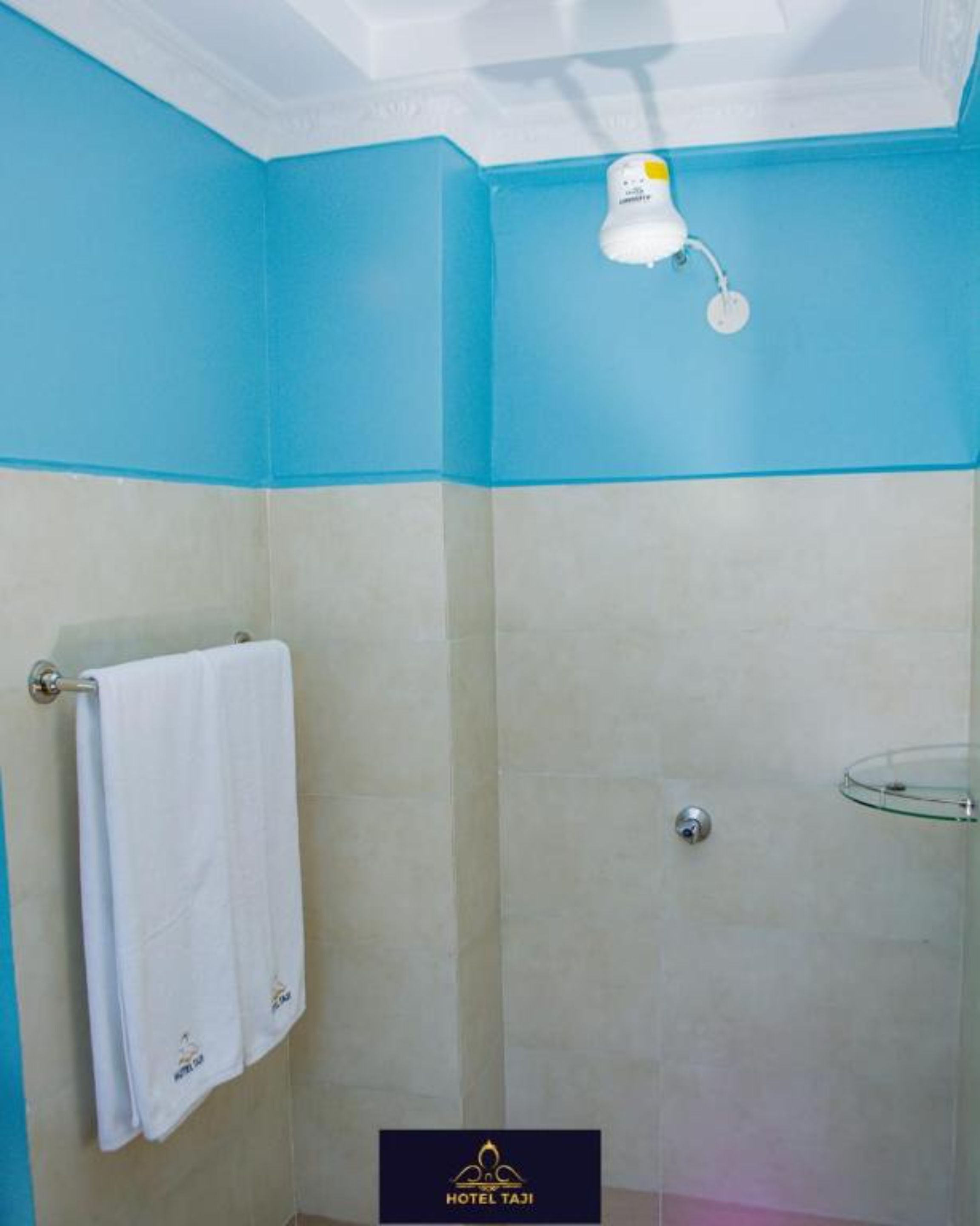 Un baño con pared azul, una toalla blanca colgada en un perchero, cerramiento de ducha de vidrio y el logo de Hotel Taji en la parte inferior.