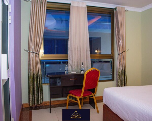 Une chambre d’hôtel avec un lit, un bureau et une chaise. Il y a une fenêtre avec des rideaux, une bouteille d’eau et un verre sur le bureau.