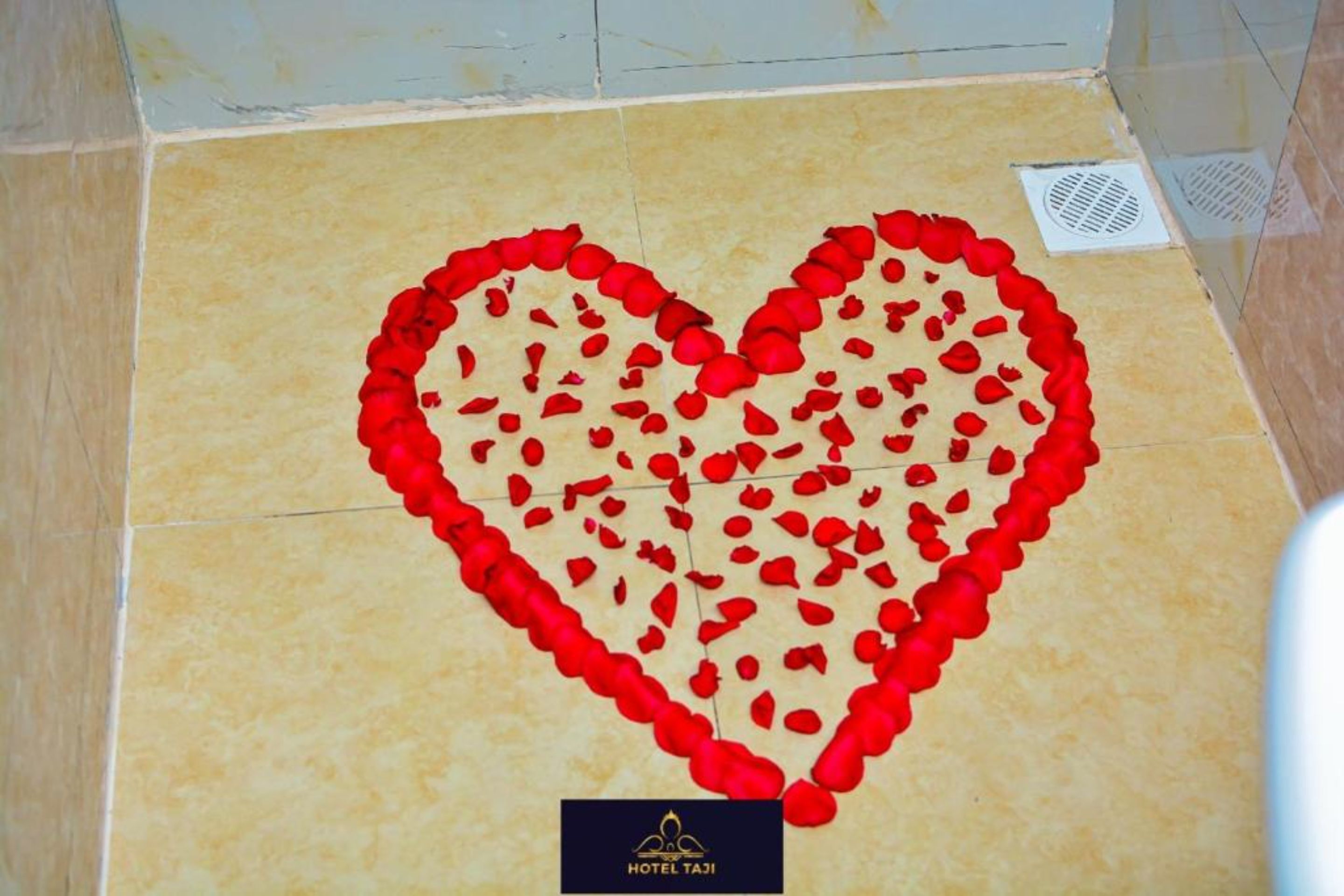 A heart-shaped decoration made of red rose petals on a tiled floor, with a Hotel Taj logo at the bottom.