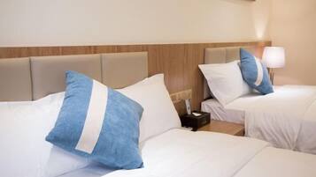 A hotel room with two beds, each with white bedding and blue pillows. There is a nightstand with a lamp and a small table between the beds.