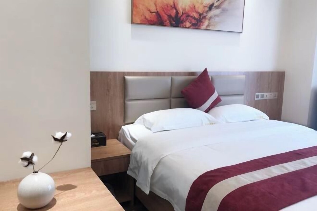 A hotel room with a bed, a nightstand, a vase with flowers, and a painting on the wall.
