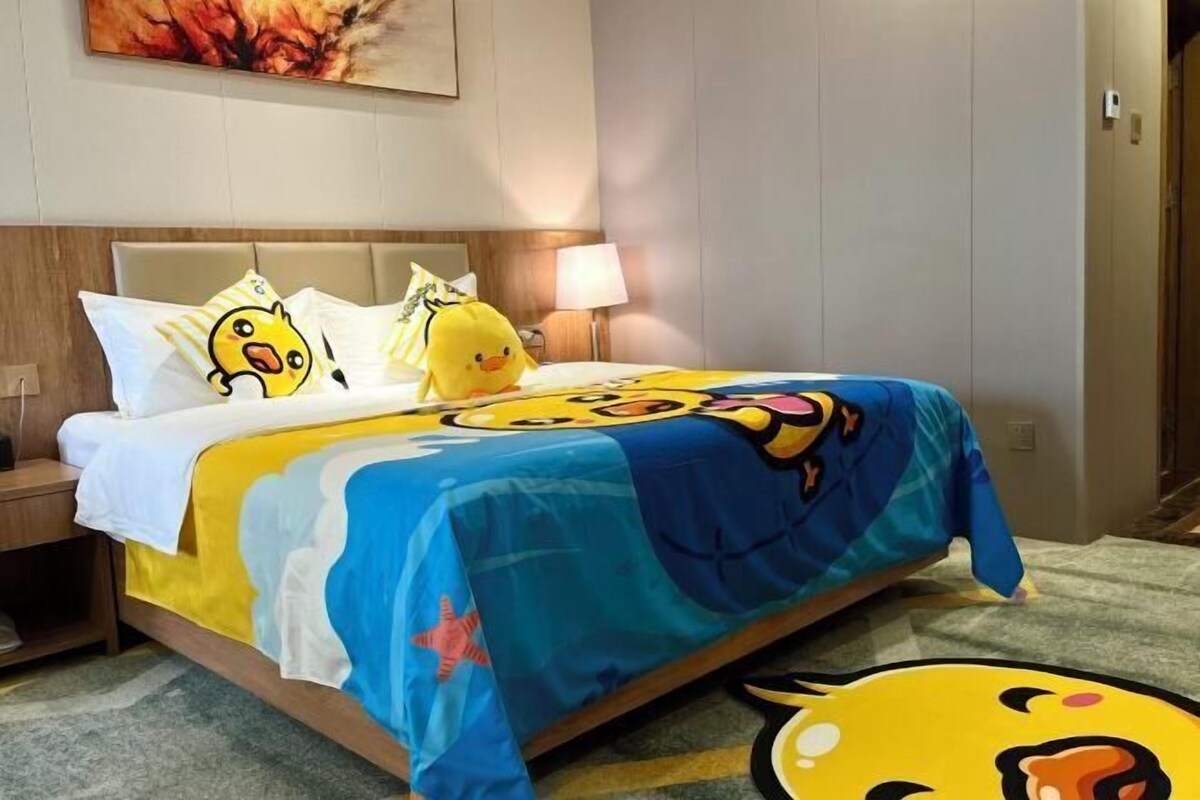 A hotel room with a bed featuring cartoon duck-themed bedding, a bedside lamp, and a wall-mounted painting.
