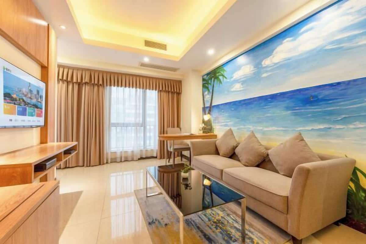 A modern living room with a beige sofa, a coffee table, and a TV on a wooden stand. A large painting of a beach scene is on the wall.