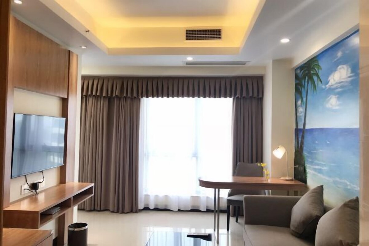 A modern living room with a flat-screen TV, a wooden entertainment unit, a glass coffee table, a sofa, a dining table with chairs, and a large window with curtains.
