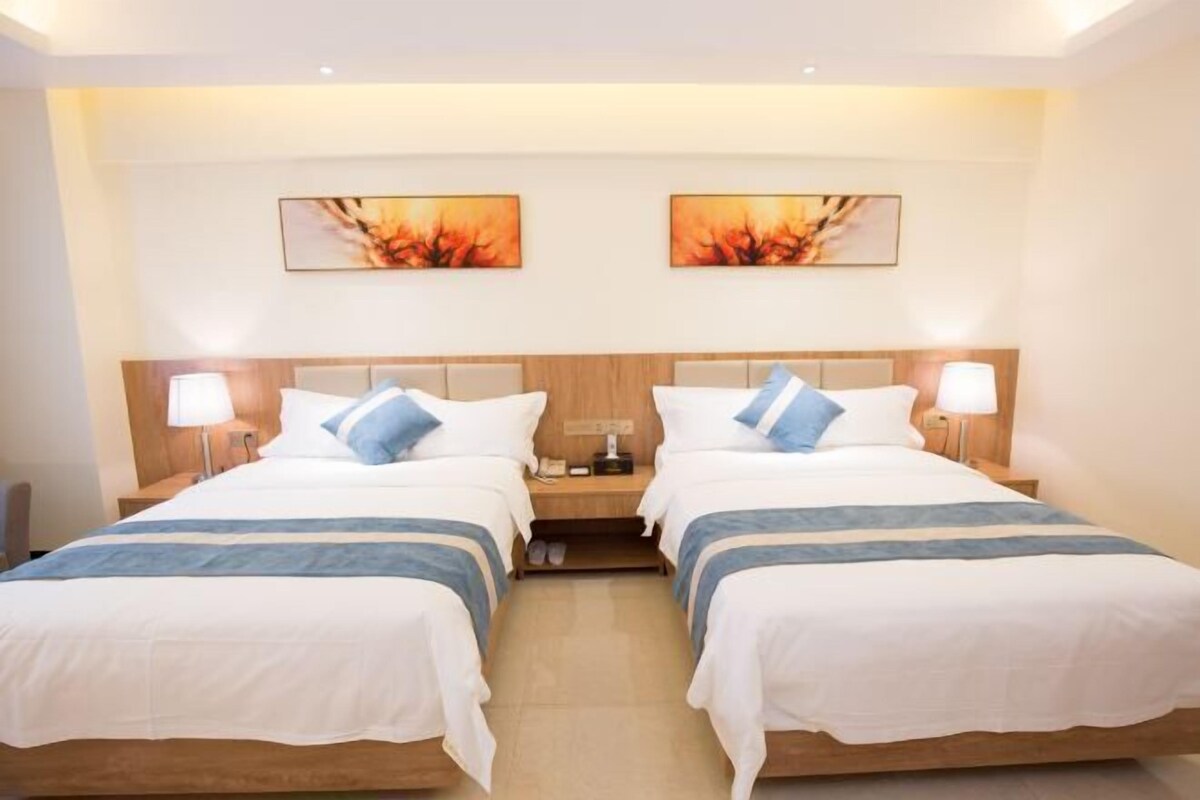 A hotel room with two beds, each with a wooden headboard, white bedding, and blue accents. There are two bedside tables with lamps, and artwork is displayed on the wall above the beds.